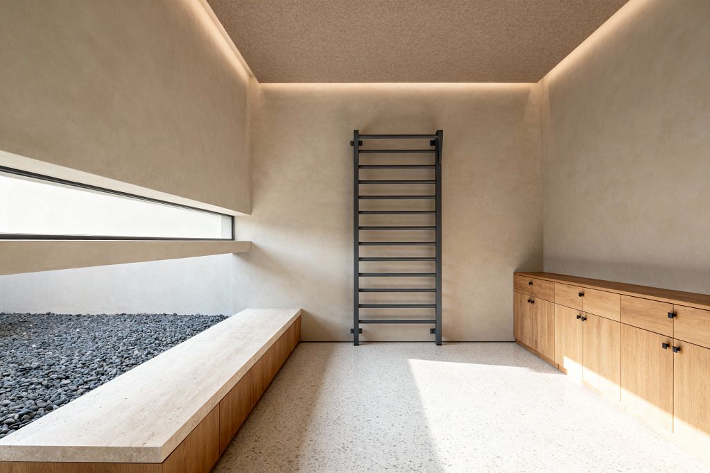 A minimalist home gym centers a black wall-mounted stall bar on a warm beige plaster wall, with a long run of natural oak cabinets with black pulls to the right and a low bench topped with light travertine beside a recessed bed of gray river pebbles to the left. A slim horizontal window and perimeter cove lighting brighten the pale terrazzo floor, emphasizing clean lines and an uncluttered layout suited to stretching and body-weight training.