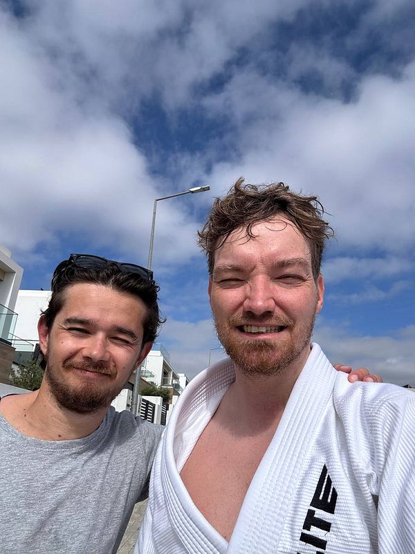 Two men pose for a selfie outdoors, one wearing a BJJ gi.