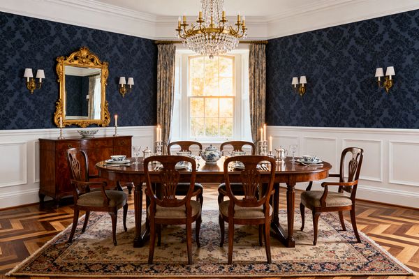 A formal dining room with navy damask wallpaper above white wainscoting centers a polished mahogany table surrounded by six carved chairs. A brass-and-crystal chandelier hangs over blue-and-white china and silver candlesticks. A gilded mirror tops a mahogany sideboard, with brass wall sconces flanking the walls. Herringbone parquet and a floral oriental rug ground the space, while pleated blue drapery frames a sunlit bay window.