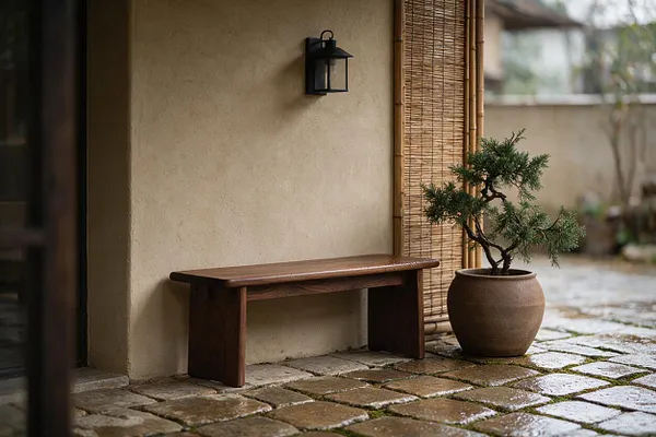 A serene Japandi-style exterior nook with a simple wooden bench against a textured beige stucco wall, a black lantern mounted above, and a potted bonsai beside a bamboo screen. The stone-paved ground adds an earthy, natural feel.