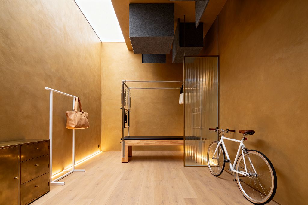 A contemporary home gym with warm ochre plaster walls and pale oak floors centers on a Pilates Cadillac with a stainless frame and wood base. Gray felt acoustic cubes hang from the ceiling near a linear skylight, while concealed LED lighting washes the lower walls. At left is a brass three-drawer cabinet and a slim white clothes rail holding a tan tote; at right a white bicycle rests beside a ribbed glass partition.