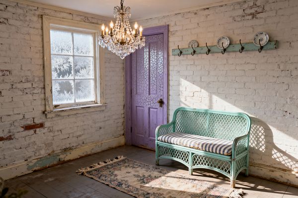 A shabby chic mudroom with whitewashed brick walls, a frosted-paned window, and a crystal chandelier. A lavender, crackle-painted wooden door sits beside an aqua peg rail with wrought-iron hooks displaying blue-and-white plates. A mint-painted rattan settee with a gray-and-cream striped cushion rests above a faded floral runner on worn wood floors, capturing a softly lit, timeworn charm.