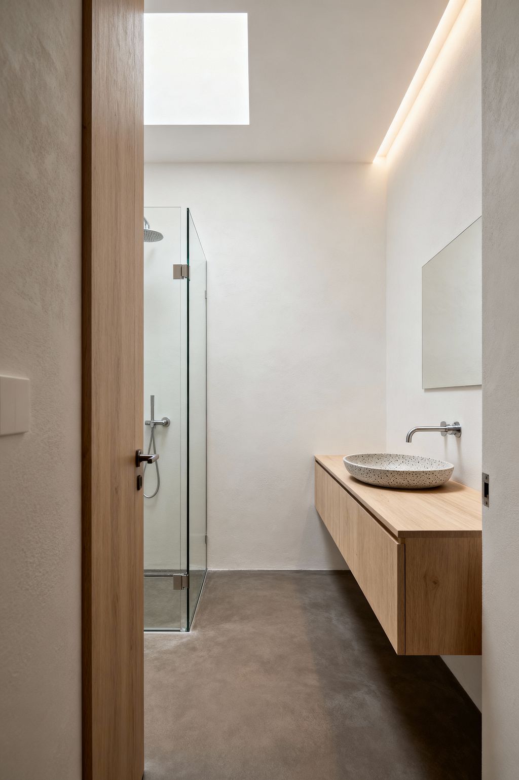 A narrow minimalist bathroom with warm white plaster walls and a large ceiling skylight, softened by concealed cove lighting. To the right, a floating light-oak vanity holds a speckled terrazzo vessel sink with a wall-mounted stainless steel faucet beneath a frameless mirror. At the end of the room, a frameless glass shower with a rain head sits on a smooth gray microcement floor, emphasizing clean lines and open space.