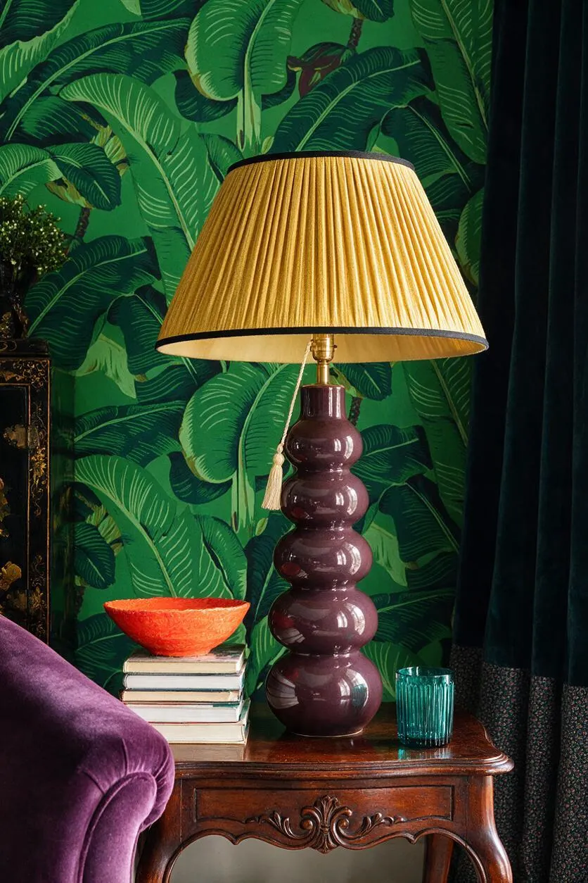 A decorative table lamp stands on a carved wooden side table against bold green tropical leaf wallpaper. The lamp has a glossy stacked purple ceramic base and a large pleated beige shade, with books and colorful glass accents nearby.