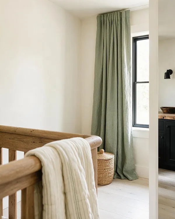 A bright farmhouse corner with creamy white walls, a tall window dressed in muted sage linen curtains, and a woven lidded basket on pale wood flooring. In the foreground, a natural oak rail is draped with a striped cream textile, while a black metal wall sconce and dark cabinetry peek in from the adjacent room.