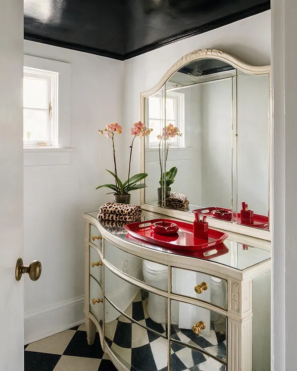 A compact powder room with a curved mirrored vanity in cream and brass, topped with glass and a bright red tray. An ornate matching mirror reflects a window, while a black ceiling and black-and-cream checkerboard floor add drama. Pink orchids and leopard-print towels reinforce the glamorous Hollywood Regency style.