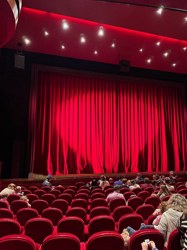 A theater interior with a red curtain and audience members seated in red chairs.