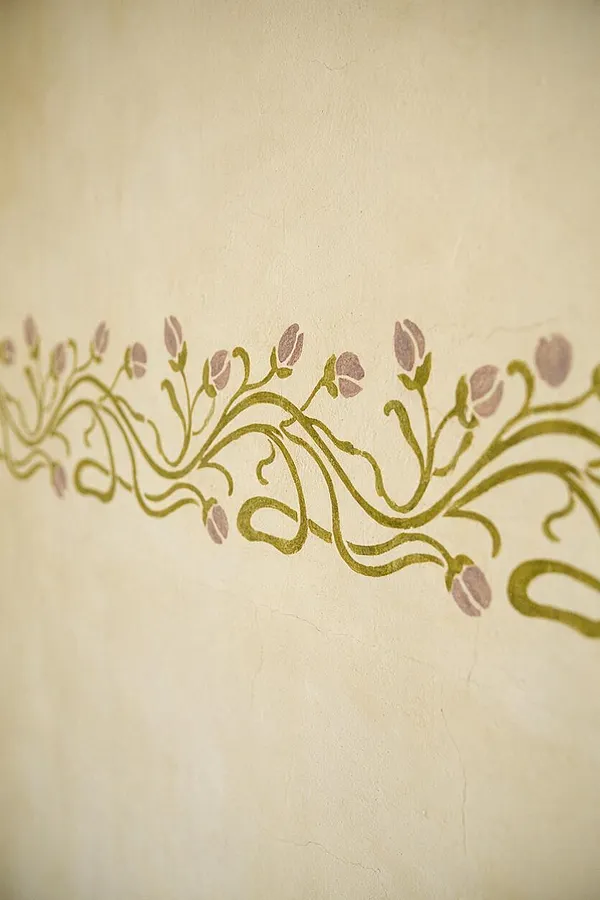 A close-up of a cream plaster wall featuring a horizontal hand-painted Art Nouveau border. The design shows flowing olive-green vine-like lines with small muted pink and lavender buds, creating a soft organic pattern across the surface.