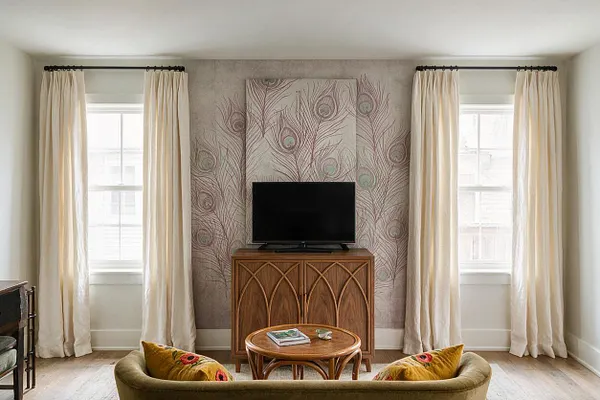 A bright media room with a centered flat-screen TV on a carved wooden cabinet, set against peacock-feather wallpaper in soft taupe and green. Cream curtains frame two tall windows, and a curved olive-green sofa with mustard pillows sits in front of a round wooden coffee table.