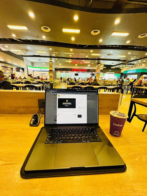 A laptop is set up on a wooden table in a bustling food court, accompanied by a drink.