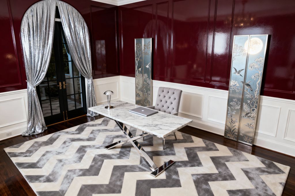 A glamorous home office centers on a white marble desk with polished chrome X-legs atop a plush gray-and-ivory chevron rug, paired with a tufted light-gray chair and a chrome dome lamp. High-gloss burgundy paneled walls above white wainscoting reflect two tall etched mirrored panels and black French doors framed by arched, shimmering silver drapery, while dark hardwood floors complete the Hollywood Regency look.