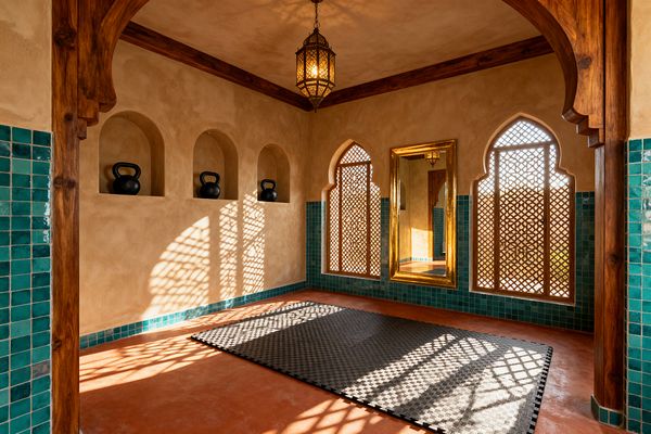 A Moroccan-inspired home gym with sandy tadelakt plaster walls, three arched niches holding black kettlebells, and a terracotta floor topped by a charcoal interlocking mat. The lower walls are clad in teal square zellige tiles, while ogee-arched windows with wooden mashrabiya screens cast patterned light. A brass pendant lantern hangs from the ceiling, a tall gilded mirror reflects the room, and carved wood arches and beams frame the space.