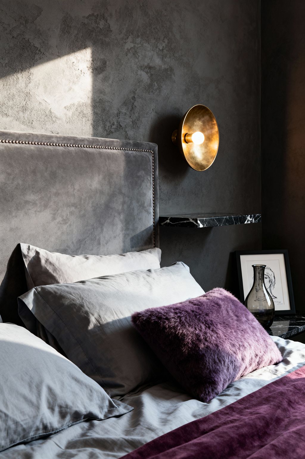 A contemporary bedroom corner features a gray velvet upholstered headboard with nailhead trim against a charcoal textured wall. A round brushed-brass sconce with an exposed bulb glows above a thin black marble floating shelf holding a smoked-glass vessel and a small framed line drawing. The bed is dressed in soft gray sheets with a deep plum velvet runner and a purple faux-fur cushion, while angled daylight casts dramatic shadows and warmth.