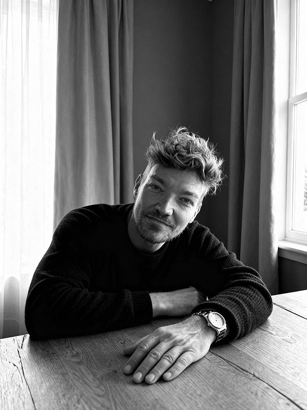 A man poses for a black and white photo shoot, resting his arms on a wooden table.