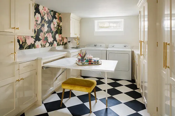 A compact basement laundry room with cream cabinetry, brass hardware, a black-and-white checkered floor, and a white folding table centered in the space. A bold floral wallpaper accent wall adds Hollywood Regency flair, while a mustard velvet stool, white appliances, and a small high window brighten the room.