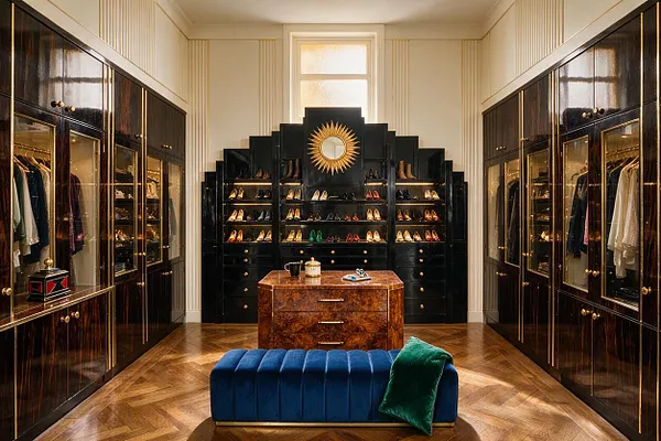 A symmetrical walk-in closet with glossy dark cabinetry, gold-trimmed glass display doors, and a stepped black back wall centered by a sunburst mirror. Clothing and shoes are neatly arranged on both sides, with a rich wood dresser, a blue velvet bench, and parquet flooring completing the elegant Art Deco look.