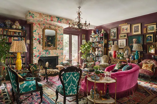 A richly decorated basement living room filled with layered patterns, framed artwork, and vintage furnishings. Floral wallpaper wraps the fireplace surround, while a hot pink curved sofa, patterned armchairs, brass lighting, and dark painted walls create a dramatic maximalist look.
