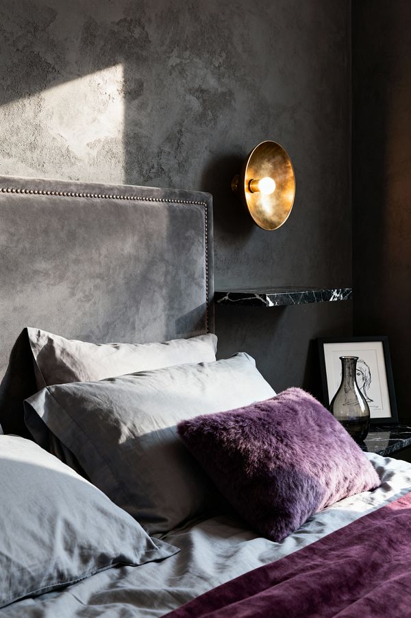 A contemporary bedroom corner features a gray velvet upholstered headboard with nailhead trim against a charcoal textured wall. A round brushed-brass sconce with an exposed bulb glows above a thin black marble floating shelf holding a smoked-glass vessel and a small framed line drawing. The bed is dressed in soft gray sheets with a deep plum velvet runner and a purple faux-fur cushion, while angled daylight casts dramatic shadows and warmth.
