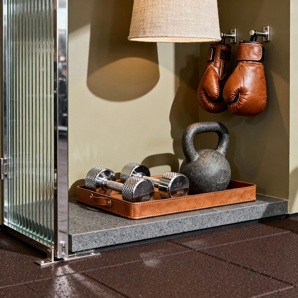 A close-up of a transitional home gym vignette: a fluted glass divider with polished chrome frame sits beside a muted olive wall lit by a linen-shaded sconce. Cognac leather boxing gloves hang from metal hooks above a gray stone ledge, where a leather tray organizes two chrome dumbbells and a cast-iron kettlebell; dark brown rubber floor tiles complete the space.