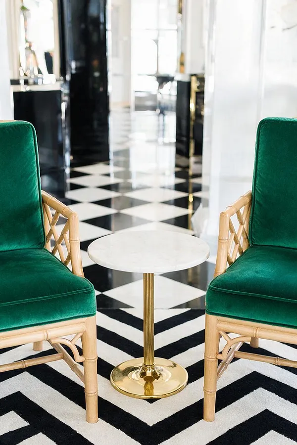 A glamorous seating vignette with two emerald green velvet chairs facing a small round white tabletop on a polished brass pedestal. The room features a bold black-and-white geometric floor and bright white walls, creating a refined Hollywood Regency look.
