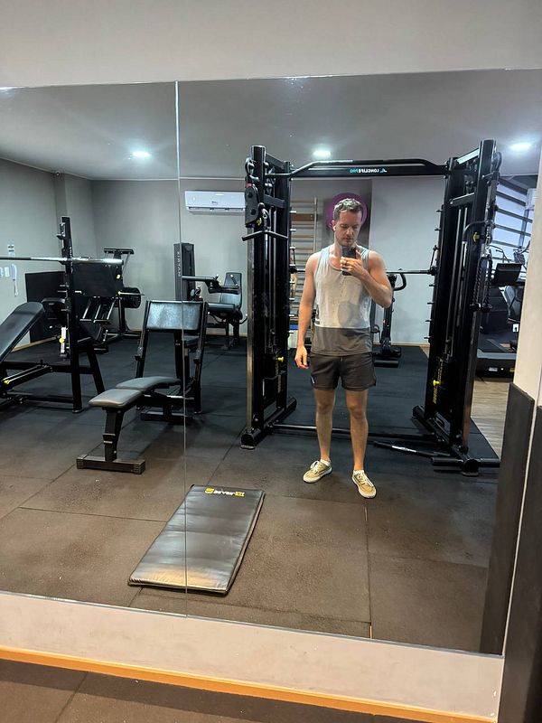A person stands in a gym, taking a selfie in the mirror while surrounded by workout equipment.