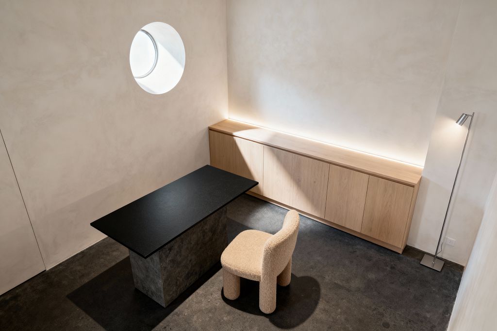 A minimalist home office with a black stone desk on a concrete pedestal set on a charcoal concrete floor. A sand-colored bouclé chair faces the desk; a long run of light oak cabinets lines the wall, lit by a recessed LED strip, while a circular window casts a beam of daylight and a slim stainless steel floor lamp stands in the corner against warm white plaster walls.