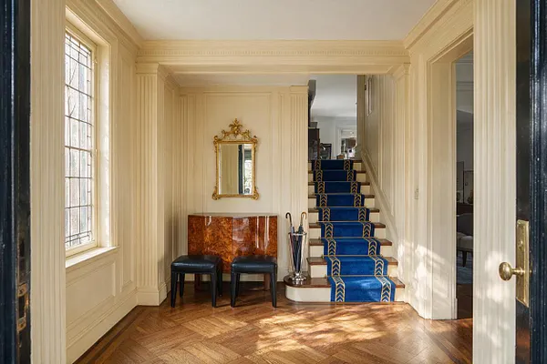 An Art Deco entryway with cream paneled walls, a herringbone wood floor, and a staircase covered in a vivid blue runner with gold geometric trim. A small lacquered console, black stools, a gold-framed mirror, and a chrome umbrella stand create a refined, classic look.