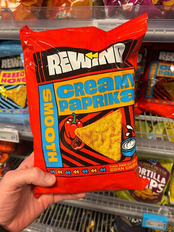A hand holds a bright red bag of creamy paprika-flavored corn chips in a grocery store aisle.