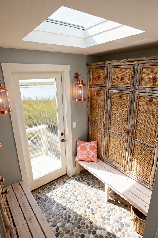 A bright coastal mudroom with a skylight and a ribbed-glass exterior door. Along the right wall, tall built-in lockers combine driftwood frames with woven rattan doors and copper knobs; matching copper lantern sconces flank the door. Light, whitewashed wood benches run along both sides, one topped with a coral-pattern pillow, and a woven basket sits below. The floor is a pebble mosaic in gray, sand, and slate tones, and the walls are soft gray with white trim.