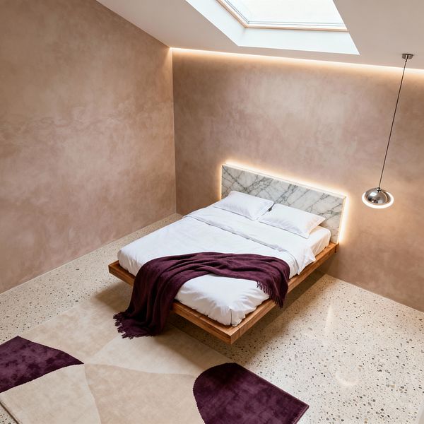 A minimalist modern bedroom with a floating oak platform bed and a white marble headboard lit by a warm LED halo. The room features warm beige plaster walls, a skylight and perimeter cove lighting, light terrazzo flooring, a cream-and-plum geometric rug, crisp white bedding layered with a deep plum throw, and a single chrome pendant hanging to the right.