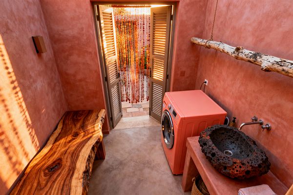 A compact bohemian laundry room with coral-pink plaster walls and a smooth concrete floor features a coral front-loading washer, a porous black lava-stone vessel sink with a wall-mounted stainless faucet, and a live-edge wood bench. A natural branch suspended by rope serves as a hanging rod. Louvered double doors open to a patio veiled by a red-orange beaded curtain, with warm sunlight casting patterned shadows across the space.