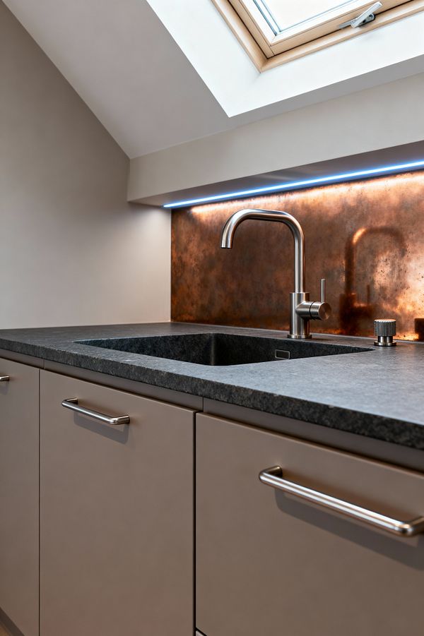 Close-up of a modern kitchen sink beneath a skylight, featuring a honed dark gray granite countertop with integrated basin, a brushed stainless gooseneck faucet, and matte taupe flat-panel cabinets with bar pulls. A patinated copper sheet backsplash glows under a slim, cool-toned LED strip set beneath a shallow shelf, while the sloped white ceiling and light wood-framed skylight bring natural light to the compact, minimalist workspace. The overall palette balances warm metal with neutral cabinetry for a clean, contemporary look.