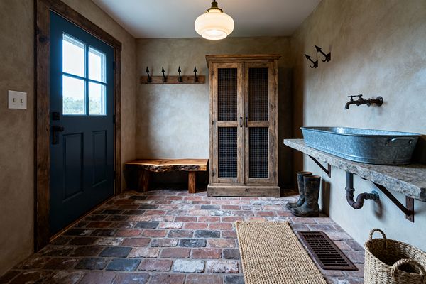 A rustic mudroom features a deep blue door framed with rough wood, greige plaster walls, and a worn brick floor. A live-edge bench and arrowhead iron hooks sit beside a tall wood cabinet with wire-mesh doors; opposite, a rough stone shelf holds a galvanized trough sink with a wall-mounted faucet and exposed pipe, while a jute rug, wicker basket, rubber boots, and a schoolhouse ceiling light add practical character.