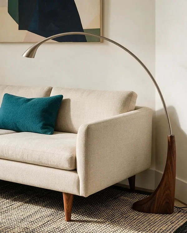 A Mid-Century Modern living room vignette with a cream upholstered sofa, a teal accent pillow, and a sweeping arc floor lamp with a wood base. The scene includes a textured woven rug, warm wood legs, and a minimalist abstract wall art piece above the sofa.