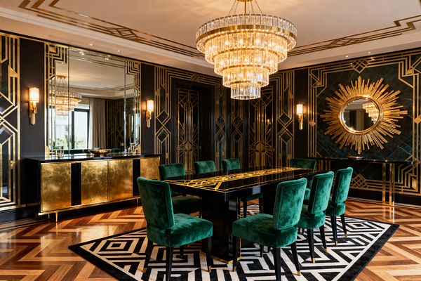 A luxurious Art Deco dining room features a black lacquer table with gold geometric inlay, surrounded by eight emerald velvet chairs atop a black-and-white graphic rug. Black walls with gold linear paneling frame mirrored sections, a large sunburst mirror, and brass sconces, while a tiered crystal chandelier, brass-clad sideboard, chevron parquet floor, and ceiling border lines create a glowing, symmetrical setting.