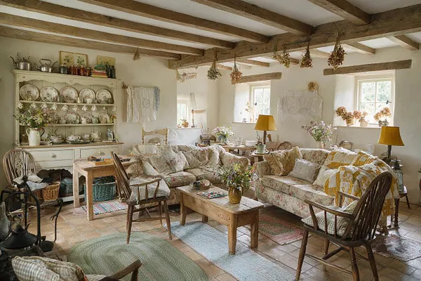 A cozy cottagecore living room with exposed wooden ceiling beams, cream plaster walls, and a mix of floral slipcovered sofas and wooden chairs. A painted cabinet displays patterned plates and teacups, while dried flowers, lace textiles, and soft yellow lamps create a warm, nostalgic atmosphere.
