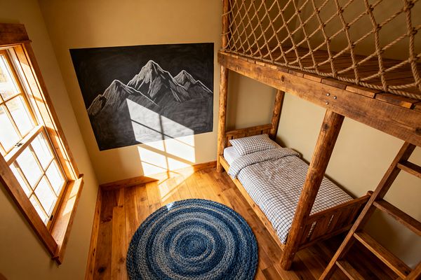 An airy, rustic kids’ bedroom features a timber loft with a jute rope safety net above a single wood bed dressed in white-and-navy grid bedding. Honey-toned plank floors and wood-trimmed windows glow in sunlight, a large black chalkboard shows hand-drawn mountain peaks, and a round braided blue rug centers the room, creating a warm cabin atmosphere with natural textures and simple handcrafted details.