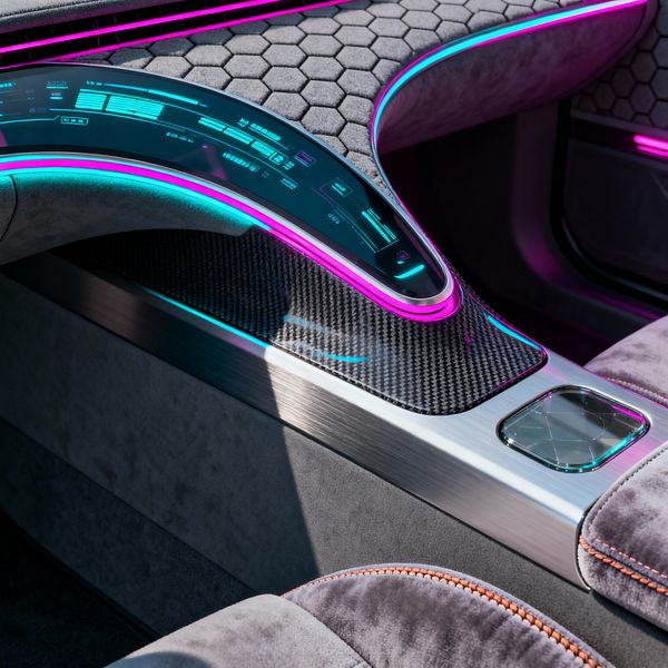 A futuristic media room control console features a sweeping curved glass touchscreen edged in neon magenta and cyan light, set above a carbon-fiber surface and a brushed aluminum surround. Gray suede-like seating with hexagonal quilting and orange topstitching frames the unit, with a square glass touchpad inset in the metal; ambient LED strips trace the contours, emphasizing smooth lines, technical textures, and an immersive high-tech atmosphere.