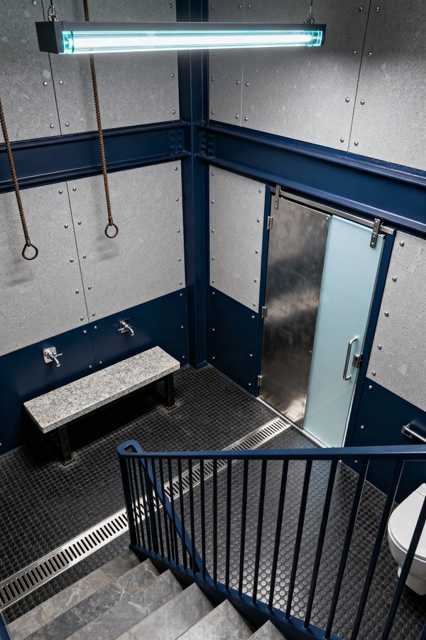 A stair leads to an industrial mudroom with black penny-round tile floors and long linear drains. Walls feature deep navy steel wainscot and riveted gray fiber-cement panels, lit by a suspended tube fixture. A granite-topped steel bench sits beneath two wall-mounted taps, and a sliding door with frosted glass and stainless hardware anchors the entry. Twisted ropes with rings hang overhead; a wall-hung toilet is partially visible.