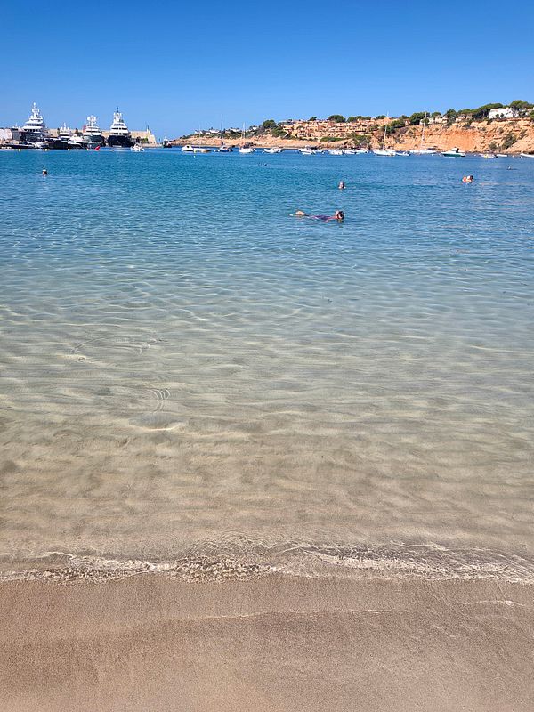 A clear beach shoreline with calm turquoise water, people swimming, and yachts docked near a rocky coastline under a bright blue sky.