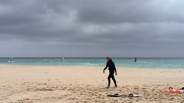 A windsurfer walks along a sandy beach with windsurfing boards in the foreground and other windsurfers in the background.