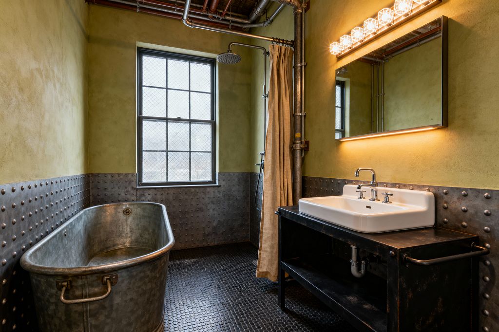 An industrial bathroom with an oval galvanized metal soaking tub set against riveted steel wainscoting and black penny-round floor tiles. Olive-toned plaster walls, an exposed rainfall shower with a canvas curtain, a wire-glass window, and a blackened steel vanity topped with a white ceramic sink sit below a wide mirror lit by warm bulbs and an LED strip, with visible copper and steel pipes throughout.