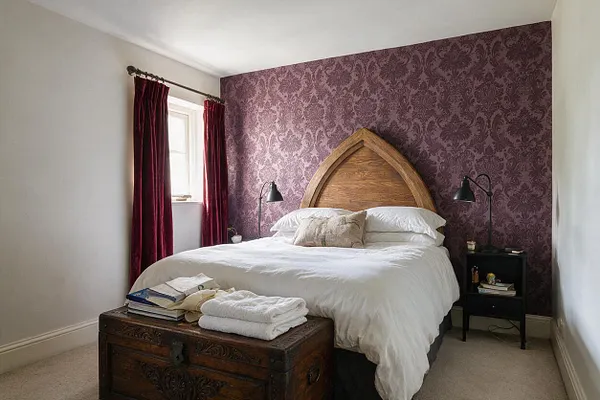 A Gothic-inspired guest bedroom with a pointed arched wooden headboard, white bedding, and dark burgundy velvet curtains. The back wall is covered in rich purple damask wallpaper, and black bedside lamps sit on either side of the bed, with an ornate dark wooden chest at the foot.