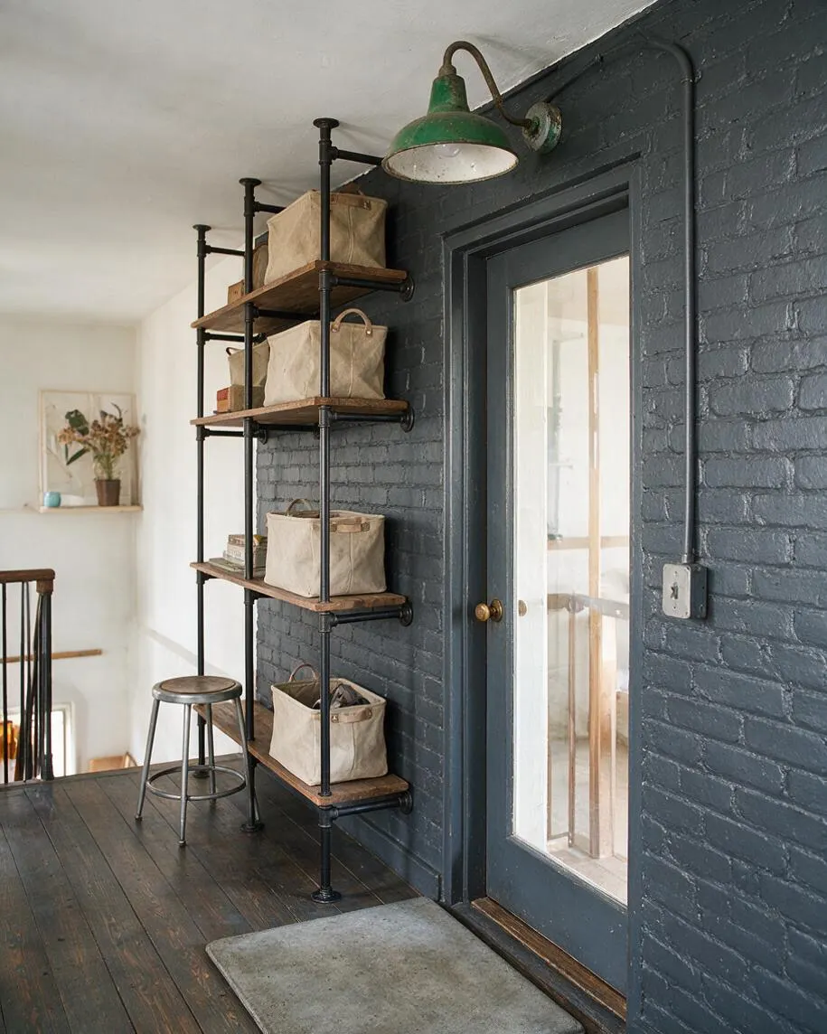 A front porch styled in industrial fashion with dark painted brick walls, a charcoal door with a large glass panel, and exposed black pipe shelving holding beige canvas storage baskets. A green metal wall sconce, wood floorboards, a metal stool, and a simple gray doormat add utilitarian charm.