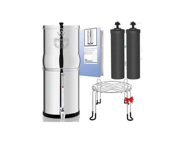 A transparent water gravity filtration system, including a glass container with a tap and a ceramic filter, sits on a patio table surrounded by greenery.