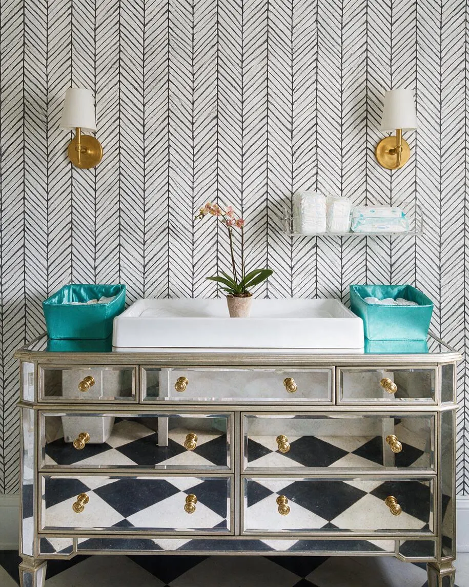 A glamorous nursery changing area with a mirrored dresser topped by a white changing pad, two teal storage bins, and a small orchid centered on top. Chevron wallpaper covers the wall behind two brass wall sconces with white shades, creating a polished Hollywood Regency look.