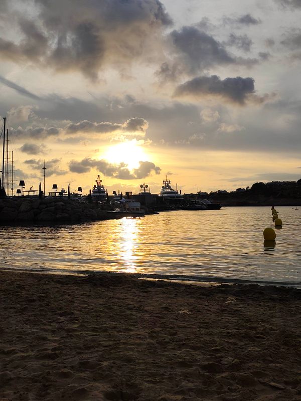 A tranquil beach scene at sunset with yachts docked near a rocky jetty and yellow buoys lining the water.