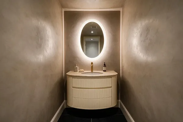 A narrow modern powder room with textured beige walls, a curved cream vanity, and a glowing oval backlit mirror centered above the sink. The space includes a light stone countertop, brass faucet, and dark floor tiles, creating a warm minimalist look.