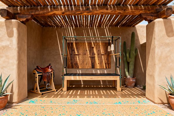 An outdoor home gym set in a stuccoed adobe alcove features a Pilates Cadillac with a patinaed metal frame and light wood base, positioned before a knotty wood pegboard wall. A Western saddle rests on a wooden stand to the left, while a tall cactus and agaves in clay pots frame the scene. Overhead vigas and latilla rafters cast striped shadows across the sand-colored walls and a concrete floor flecked with turquoise chips.