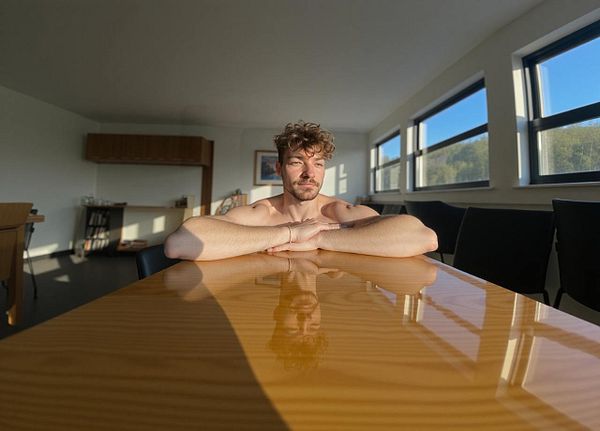 A man sits at a wooden table in a well-lit room, with sunlight streaming through large windows.